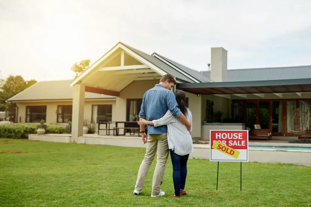 Couple standing in the front yard of the home they are selling, with a for sale sign on the lawn
